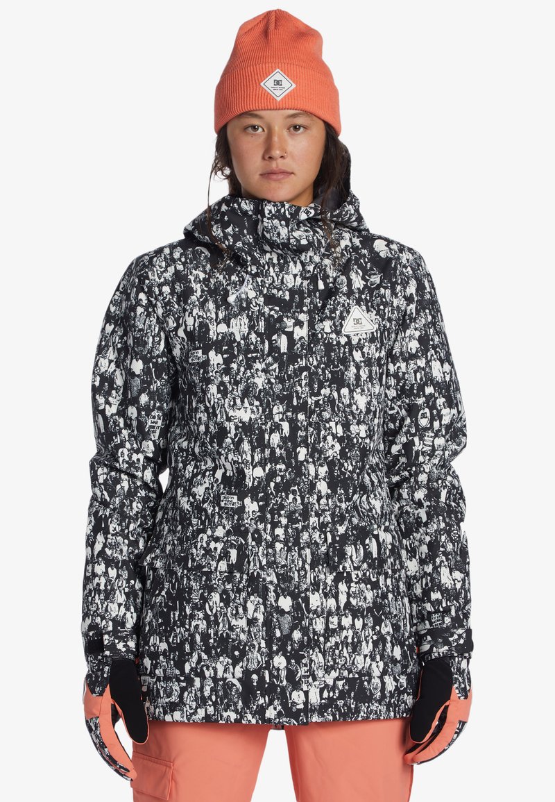 Musta-valkoinen kuvioitu eristetty takki korkealla kauluksella, yhdistetty oranssiin neulottuun pipoon ja mustiin hanskoihin, joka on päällä henkilöllä.