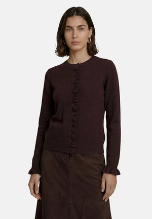 Femme aux cheveux foncés mi-longs porte un cardigan marron foncé boutonné avec des bords à volants et une jupe assortie sur un fond uni.