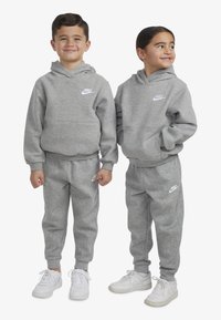 Set cu hanorac gri cu glugă și pantaloni de trening, cu un mic logo alb Nike. Materialul este moale și are un finisaj texturat, completat de adidași albi.