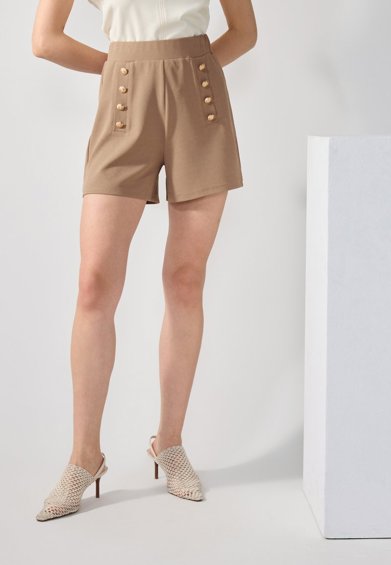 Shorts marron taille haute avec boutons sur le devant, présentant une texture lisse et des poches latérales, associés à des mules blanches tressées à bout ouvert.