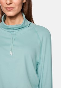 Light teal pullover with a cowl neck, featuring drawstrings and raglan sleeves. Smooth texture with a relaxed fit and minimal stitching details.