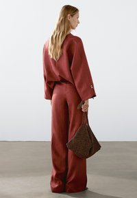 Haut ample en lin rouge avec manches larges et pantalon assorti à jambes larges. Sac à main marron texturé avec une anse nouée. Design simple, couleurs unies.