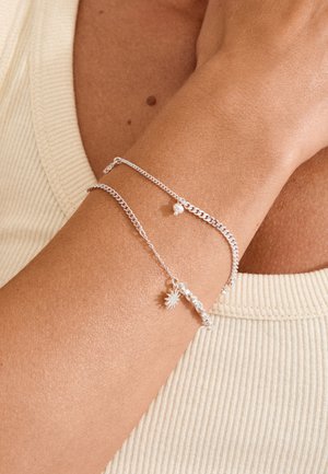 Close-up af håndled iført to delikate sølvkædearmbånd med små perle- og solformede vedhæng på hudfarvet ribstrikket top.