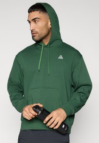 Πράσινο φούτερ με κουκούλα από απαλό ύφασμα, με μπροστινή τσέπη, κορδόνια και μικρό λευκό λογότυπο στο στήθος. Μαύρο παγούρι νερού κρατημένο στο χέρι.