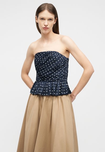 Γυναίκα που φοράει μπλε ναυτικό τοπ με πουά χωρίς τιράντες και τελείωμα με βολάν, μαζί με μπεζ πλισέ φούστα ψηλόμεση, στέκεται με τα χέρια πίσω από την πλάτη.