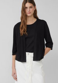 Zwarte cardigan met driekwartmouwen, open voorkant, gedragen over een zwarte top; gecombineerd met een witte rok met trekkoorddetails.