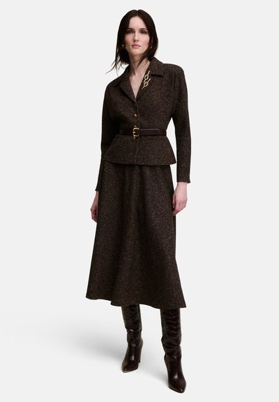 Femme portant une veste ceinturée texturée marron foncé, une jupe midi assortie et des bottes marron foncé montant aux genoux, debout devant un fond uni.