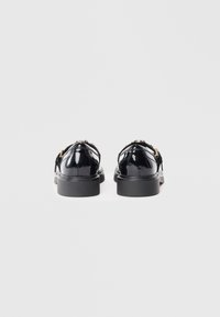 Black patent leather shoes with a rounded shape, gold hardware accents, and a thick black sole, viewed from the back.