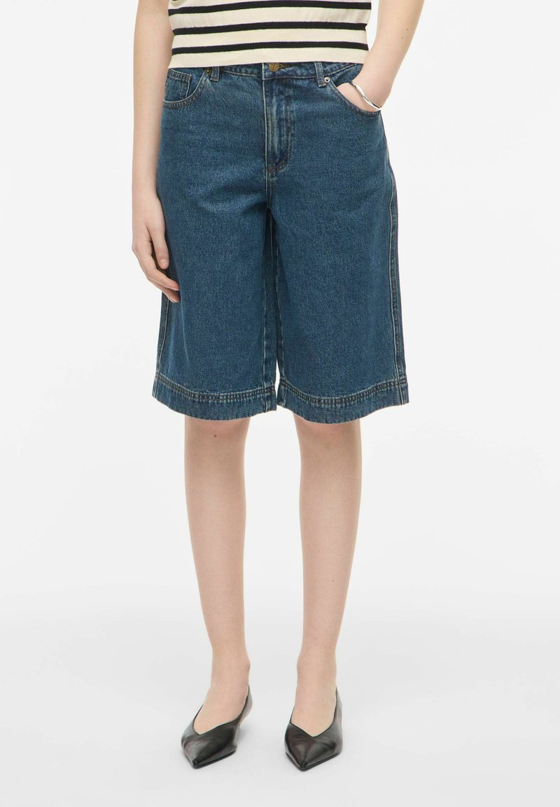Personne portant un short en jean longueur genou, un haut rayé noir et blanc et des ballerines noires à bout pointu, mains dans les poches.