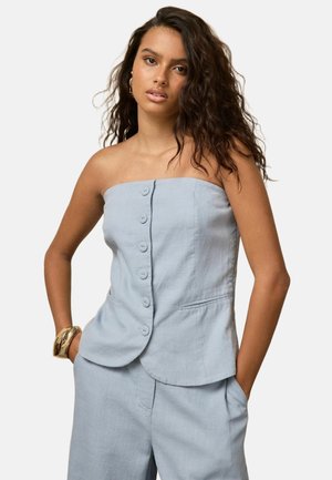 Frau mit welligem Haar, die ein trägerloses hellblaues Oberteil mit Knöpfen und passende Hose trägt, posiert mit den Händen in den Taschen und einem goldenen Armband.