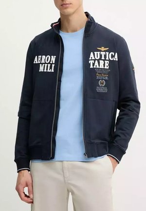 Giacca navy con zip e testo bianco, dotata di colletto alto, polsini a coste e tasche. Indossata sopra una maglietta leggera blu e pantaloni chiari.