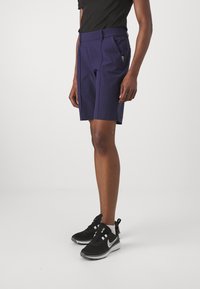 Bermude personalizate de culoare bleumarin, cu un front plat, două buzunare laterale și o textură fină. Asortate cu adidași sport negri.