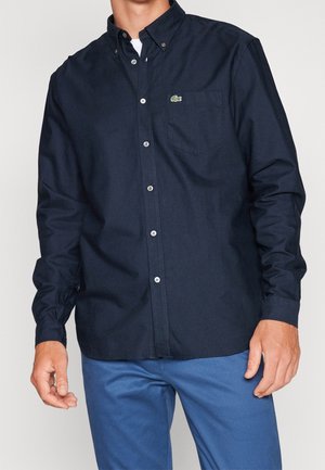 Homme portant une chemise boutonnée bleu marine foncé avec un petit logo vert sur la poche poitrine et un pantalon bleu.