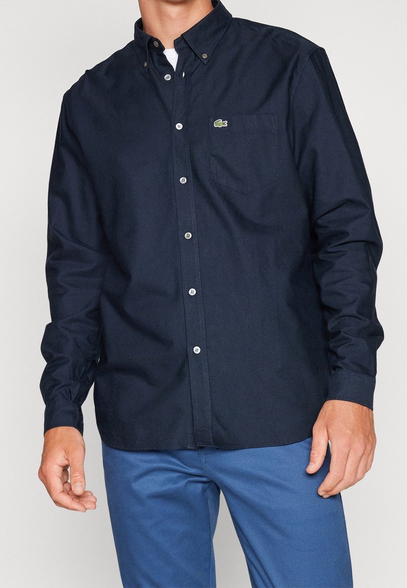 Homme portant une chemise boutonnée bleu marine foncé avec un petit logo vert sur la poche poitrine et un pantalon bleu.