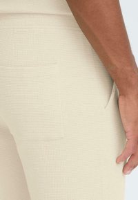Pantalon beige texturé avec un motif discret en grille, doté de poches latérales et d'une silhouette ajustée. Le tissu semble confortable et respirant.