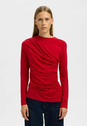 Jeune femme avec des cheveux blonds raides portant un haut rouge à manches longues froncé et un jean bleu foncé, se tenant devant un fond blanc uni.