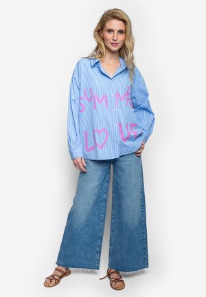 Vrouw met lang blond haar die een blauw gestreept shirt met roze tekst draagt, wijde jeans en bruine sandalen met bandjes, tegen een witte achtergrond.
