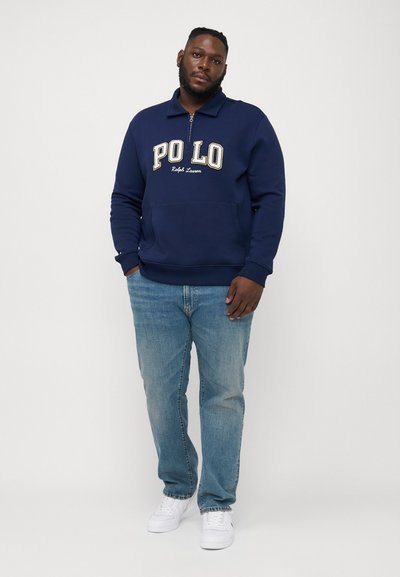 Marineblauwe zip-up sweatshirt met het "POLO"-logo in beige, gecombineerd met lichtblauwe jeans en witte sneakers. Bevat een kraag en een voorzak.