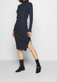 Abito a maglia blu navy, con maniche lunghe e collo alto, fessura laterale e una leggera lucentezza. Abbinato a stivaletti alla caviglia neri con stampa cocco.