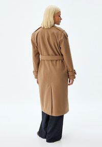 Brun ullblandad trenchcoat med axelklapp, bälte i midjan och ryggspringa. Bärs över marinblå rutiga byxor, slät textur.