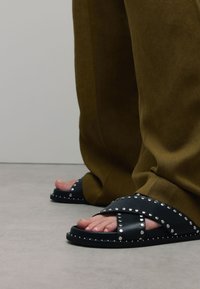 Nu-pieds noirs avec des lanières croisées, accents en clous argentés, semelles plates et orteils découverts. Associés à un pantalon ample marron foncé.