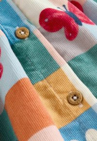 Tissu en velours côtelé multicolore avec un motif patchwork comportant des carrés roses, teal, orange et jaunes. Bouton marron avec un détail de texte "NEXT".