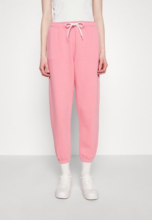 Personne portant un pantalon de survêtement rose clair avec un cordon blanc, des chaussettes blanches, des baskets blanches et un haut blanc sans manches, debout sur un sol gris.