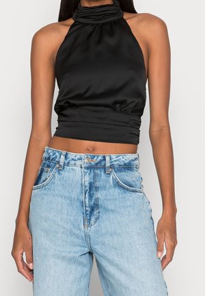 Kvinde iført en ærmeløs sort satin haltertop kombineret med højtaljede lyseblå denimjeans mod en ensfarvet baggrund.