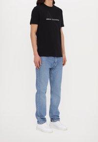 Svart bomull t-shirt med "ARMANI EXCHANGE" logotyp i vitt, kombinerat med ljusblå jeans och vita sneakers; avslappnad passform och minimalistisk design.