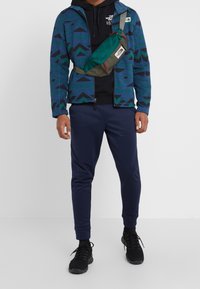 Giacca in pile blu a fantasia con zip, felpa nera con cappuccio, pantaloni sportivi blu navy e sneakers nere, accompagnati da una borsa a tracolla verde con dettagli rossi.