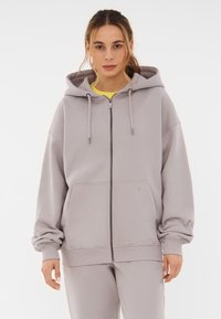 Svetlošedá mikina na zips s kapucňou, prednými vreckami a rebrovanými manžetami. Vyrobená z jemnej tkaniny s voľnejším strihom a minimalistickým dizajnom.