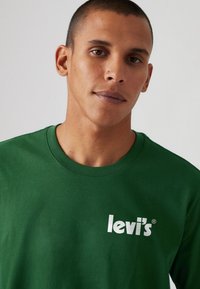 Tricou din bumbac verde cu guler rotund și mâneci scurte, având un logo alb "levi's" imprimat pe partea stângă a pieptului.