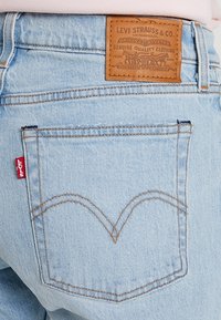 Jean en denim bleu clair avec une patch en cuir marron portant le logo de la marque et une étiquette rouge sur la poche arrière. Détail de couture courbe distinctive.