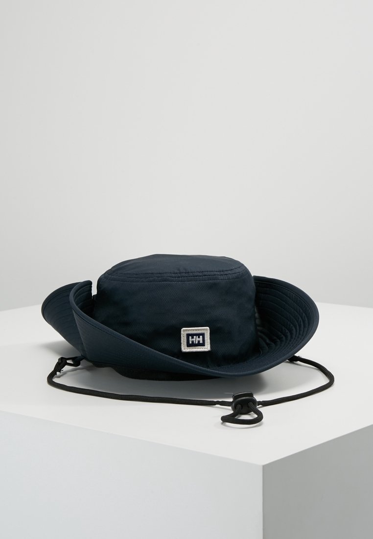 Chapeau d'extérieur à larges bords bleu marine avec sangle de menton réglable et écusson logo Helly Hansen à l'avant, posé sur une surface blanche.
