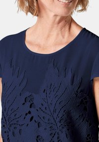 Donkerblauwe blouse met een bloemen uitsnede ontwerp aan de voorkant, gemaakt van zachte stof met een ronde halslijn en korte mouwen.