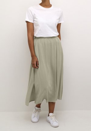 Femme portant un t-shirt blanc à manches courtes, une jupe midi vert clair avec une fente latérale, et des baskets blanches, debout devant un fond uni.