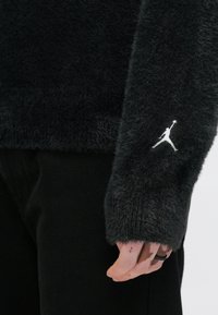 Μαύρο, χνουδωτό πουλόβερ με ριμπ μανσέτα, που διαθέτει λευκό κεντημένο λογότυπο Jumpman στο μανίκι. Φορεμένο πάνω από σκούρο παντελόνι.