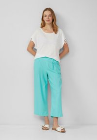 Top bianco in lino con maniche corte e lacci, abbinato a pantaloni turchesi larghi e sandali bianchi con un accento dorato.