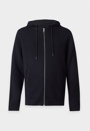 Junger Mann mit schwarzer Kapuzenjacke mit Reißverschluss, schwarzen Hosen und schwarzen Schuhen, stehend vor einem einfachen hellen Hintergrund.
