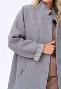 Manteau en mélange de laine grise avec un col montant, coupe décontractée et poignets à carreaux. Doté d'une fermeture à boutons et de poches latérales.