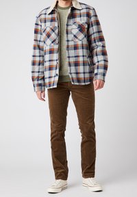 Man som bär vita sneakers, bruna kordflätade byxor, grön stickad tröja och en blå, grå, orange rutiga jacka med vit fleecekrage.