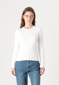 Pull en maille blanc à col cable avec des poignets et un ourlet côtelés, arborant un petit logo sur la poitrine ; associé à un jean bleu.