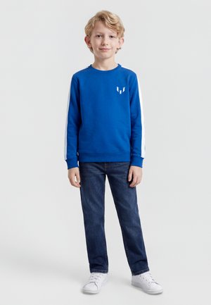 Ragazzo giovane in piedi, indossa una felpa blu con strisce bianche sulle maniche, jeans blu scuro e scarpe da ginnastica bianche, su uno sfondo semplice.