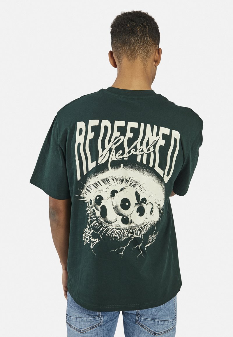 Mann trägt ein dunkelgrünes T-Shirt mit großem Grafikdruck von mehreren Augen und dem Wort "REDEFINED" auf dem Rücken, kombiniert mit hellblauen Jeans.