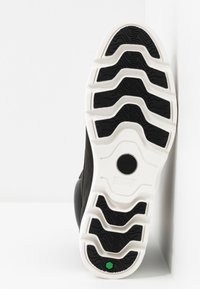 Semelle d'une chaussure de sport noire et blanche avec un grip texturé, présentant des rainures noires ondulées et un petit détail hexagonal vert près du talon.