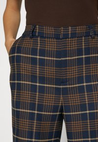 Tummansininen ruudullinen housu, jossa on tekstuurinen kangas ruskeilla ja kreeminvärisillä viivoilla, sivutaskut ja muotoiltu vyötärö.