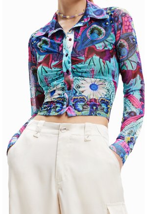 Chemise ajustée à manches longues avec motif floral abstrait vibrant et plumes de paon, associée à un pantalon beige taille haute et un collier de perles.