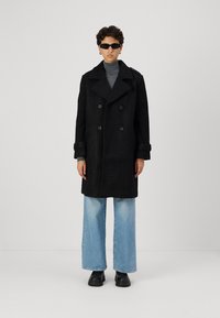 Melna, dubultā izgriezuma vilnas mētelis ar pogu aizdari. Modelis valkā pelēku turtleneck, gaiši zilas plata kāju bikses un melnas kurpes.