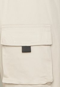 Heller beige Stoff mit einer großen aufgenähten Tasche, die über einen schwarzen Zugriemen verfügt und genähte Kanten aufweist, die eine glatte, langlebige Textur zeigen.