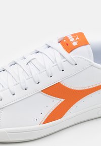 Fehér sneaker texturált narancssárga díszítéssel és kontrasztos varrással. Lapított fűzőkkel, perforációkkal az oldalon és narancssárga logócímkével rendelkezik.